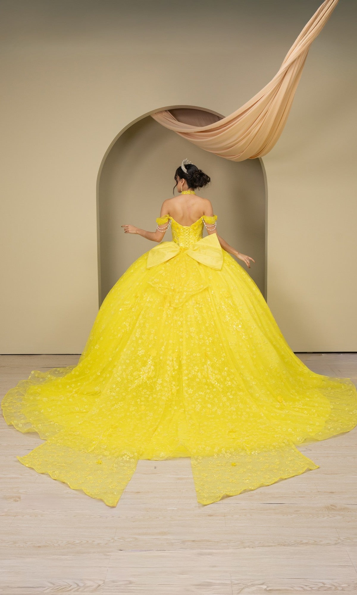 A woman wearing the Quinceanera Dress 8415JQ By Cinderella Couture, an elaborate yellow off-the-shoulder gown, stands with her back to the camera before an arched doorway on a light wooden floor.